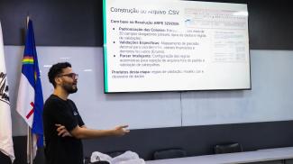 Pessoa apresenta uma palestra diante de uma tela com informações sobre a construção de arquivos CSV, enquanto gesticula para o público.